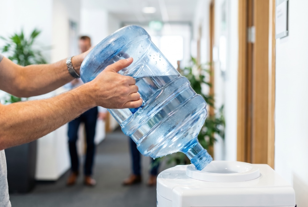 Person lifting 5 gallon water jug into an office water cooler. Indicating the inconvenience of heavy water jugs.